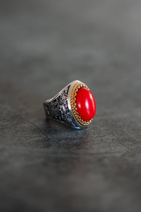 The Burning Rose Ring by Relentless Betrayal features ornate floral engravings and a large oval red stone in a gold bezel, showcased on a gray surface.