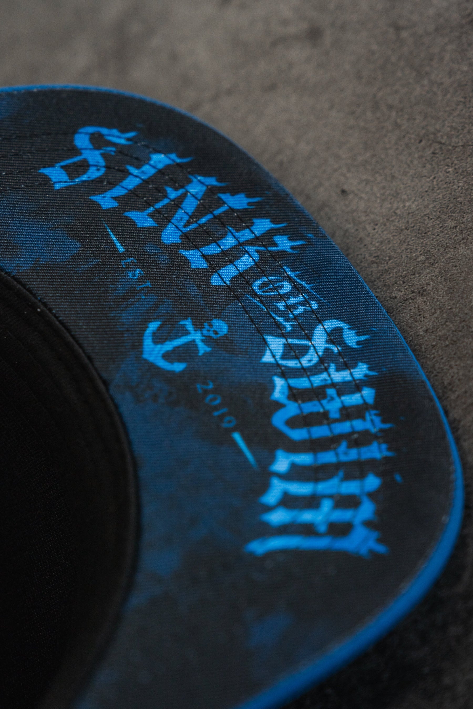 Close-up of the underside of the Relentless Betrayal Dark Waters SOS Mute PVC Snapback, featuring blue and black water-resistant brim with "SINK SWIM" lettering, an anchor graphic, and EST. 2019 printed on the surface.