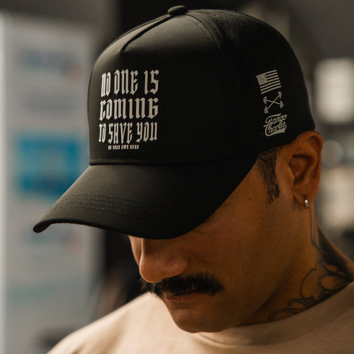 A tattooed person with a mustache looks down, wearing the Tango Charlie Apparel "No One Is Coming To Save You Hydro" snapback hat. The black cap features white gothic text, a small American flag, and additional side graphics.
