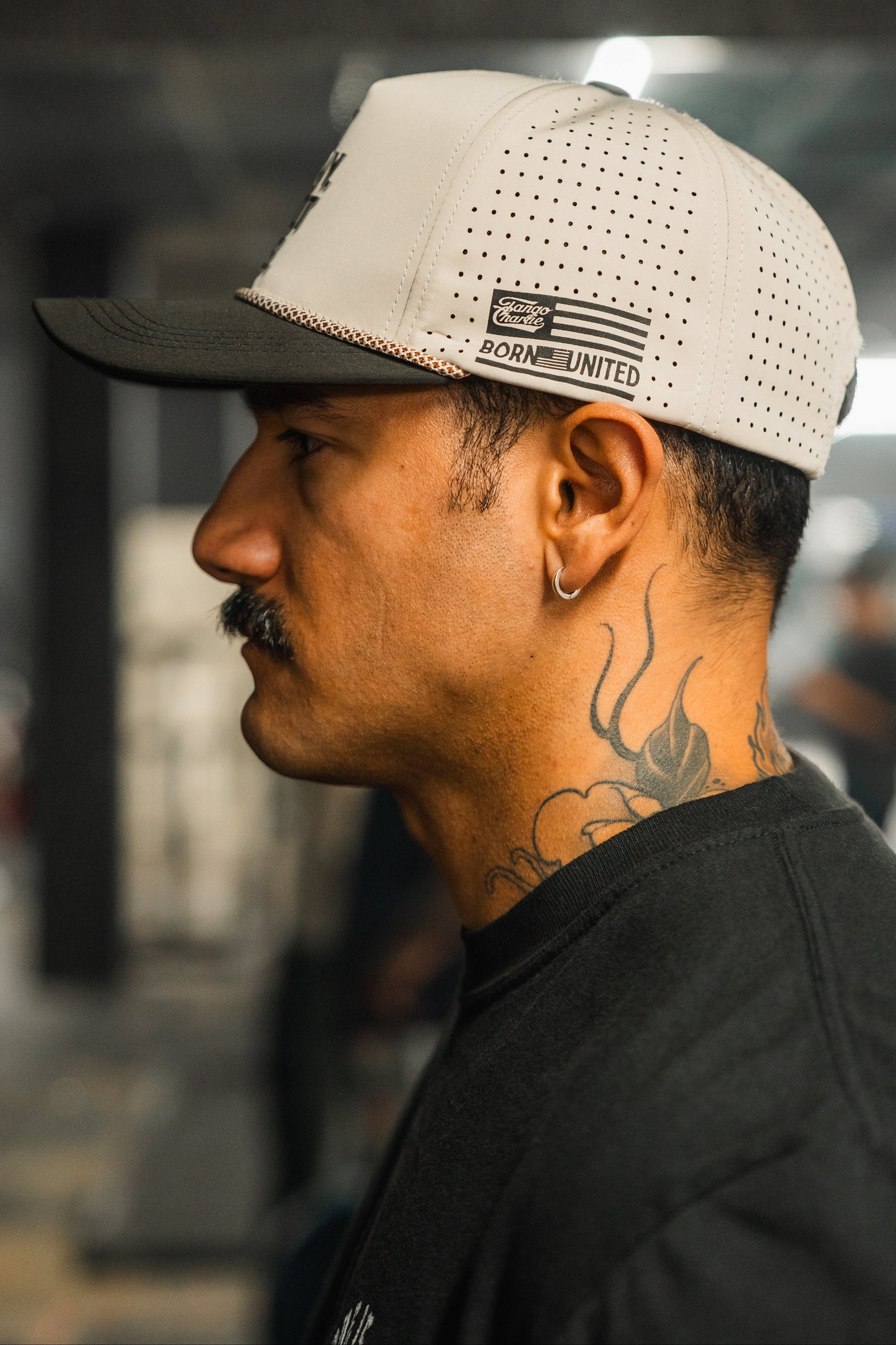 A serious-looking man with a mustache, neck tattoo, and earring is shown in profile wearing a black shirt and the white LHSG Hydro - Snapback Hat by Tango Charlie Apparel. The background is blurred.