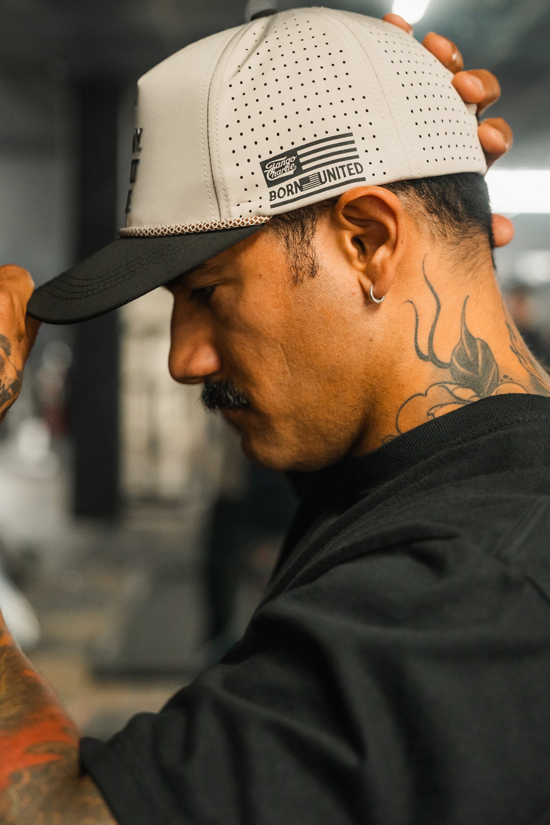A man with tattoos and a mustache adjusts his LHSG Hydro - Snapback Hat by Tango Charlie Apparel while looking down. He wears a black shirt, displaying a large floral neck tattoo, against a softly blurred indoor background.