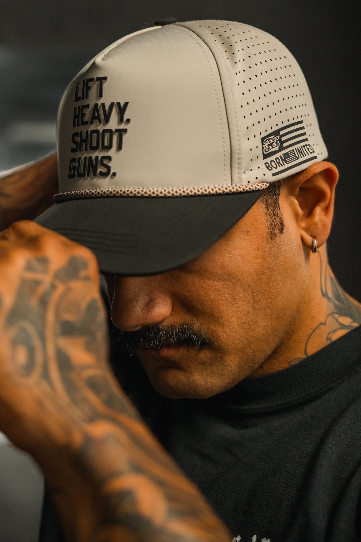 A tattooed man with a mustache adjusts a Tango Charlie Apparel LHSG Hydro Snapback Hat featuring “LIFT HEAVY, SHOOT GUNS” on the front and an American flag patch on the side, his face partially shadowed.