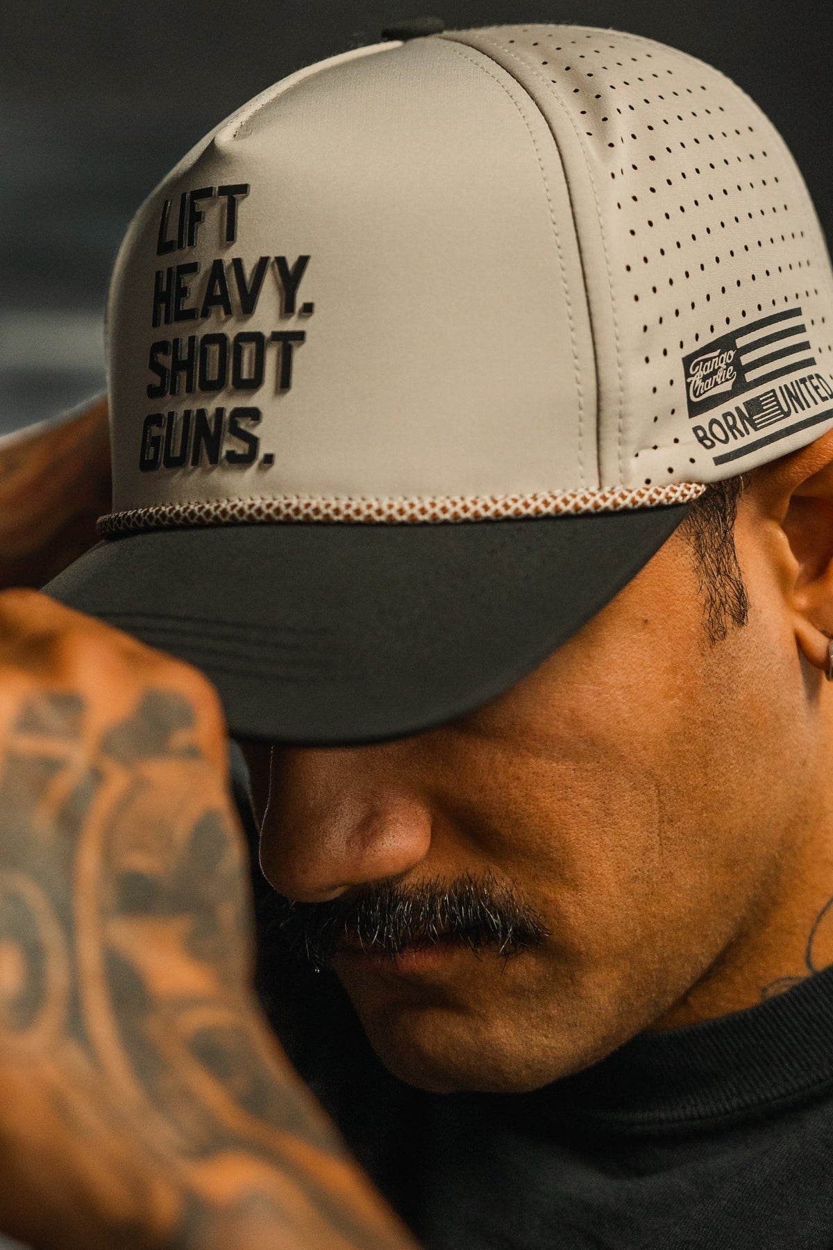 A tattooed man with a mustache adjusts the brim of his gray and black LHSG Hydro Snapback Hat by Tango Charlie Apparel, featuring “LIFT HEAVY, SHOOT GUNS” from the Born United collection.