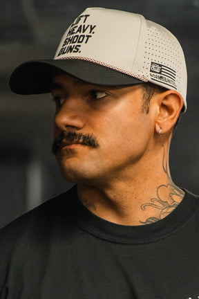 A man with a mustache, hoop earring, and neck tattoo looks to the side in a dimly lit setting wearing a Tango Charlie Apparel LHSG Hydro Snapback Hat with text and a dark shirt.