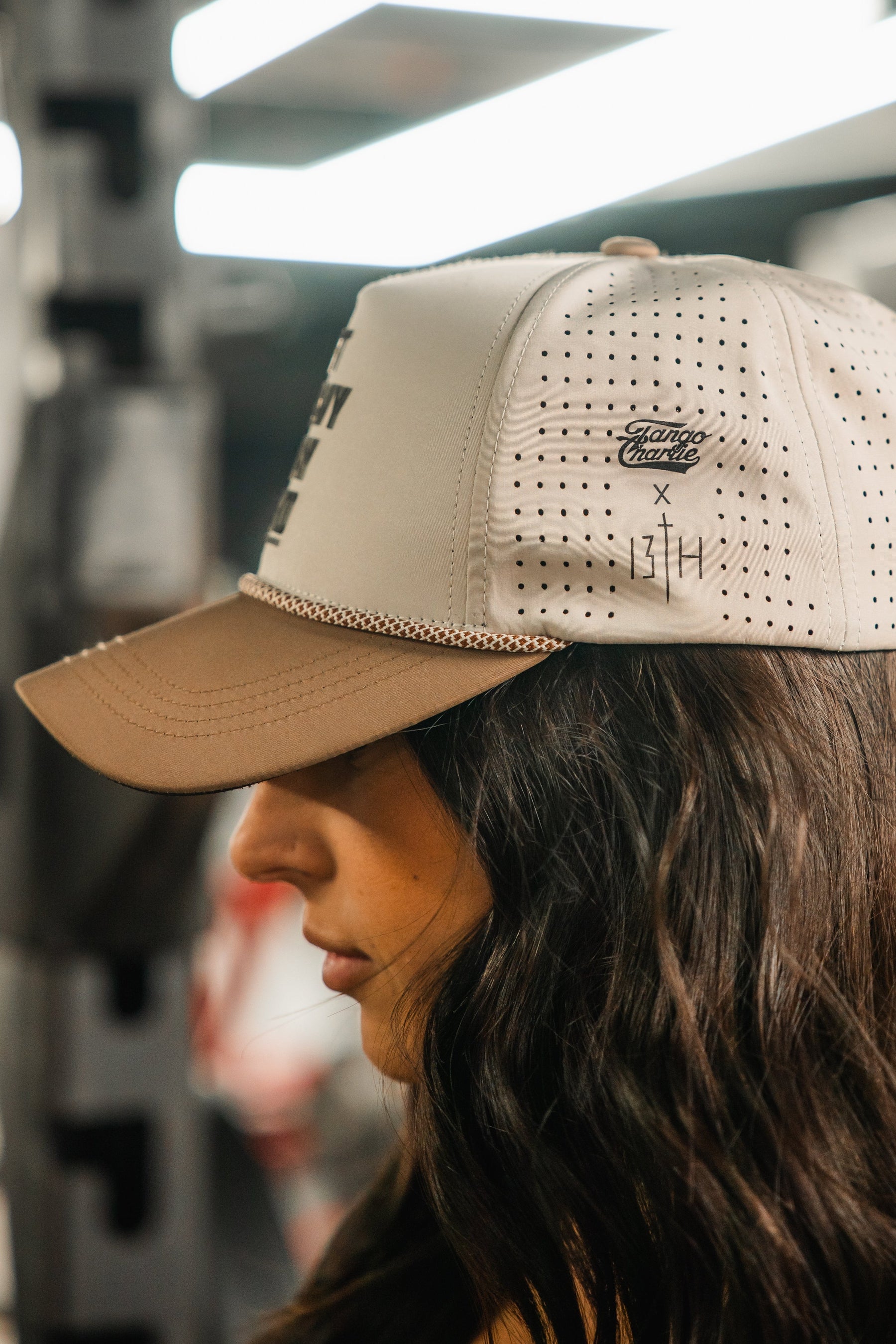A woman with long dark hair wears the Tango Charlie Apparel "Lift Heavy. Pray Hard. Hydro - Snapback Hat," a beige water-resistant cap with side ventilation holes and logos, shown in focus against a blurred background.