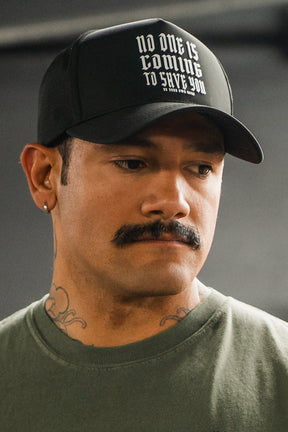 A man with a mustache and neck tattoos wears the black "No One Is Coming To Save You Hydro" snapback hat by Tango Charlie Apparel. He pairs it with a green shirt and looks slightly downward, serious-faced.