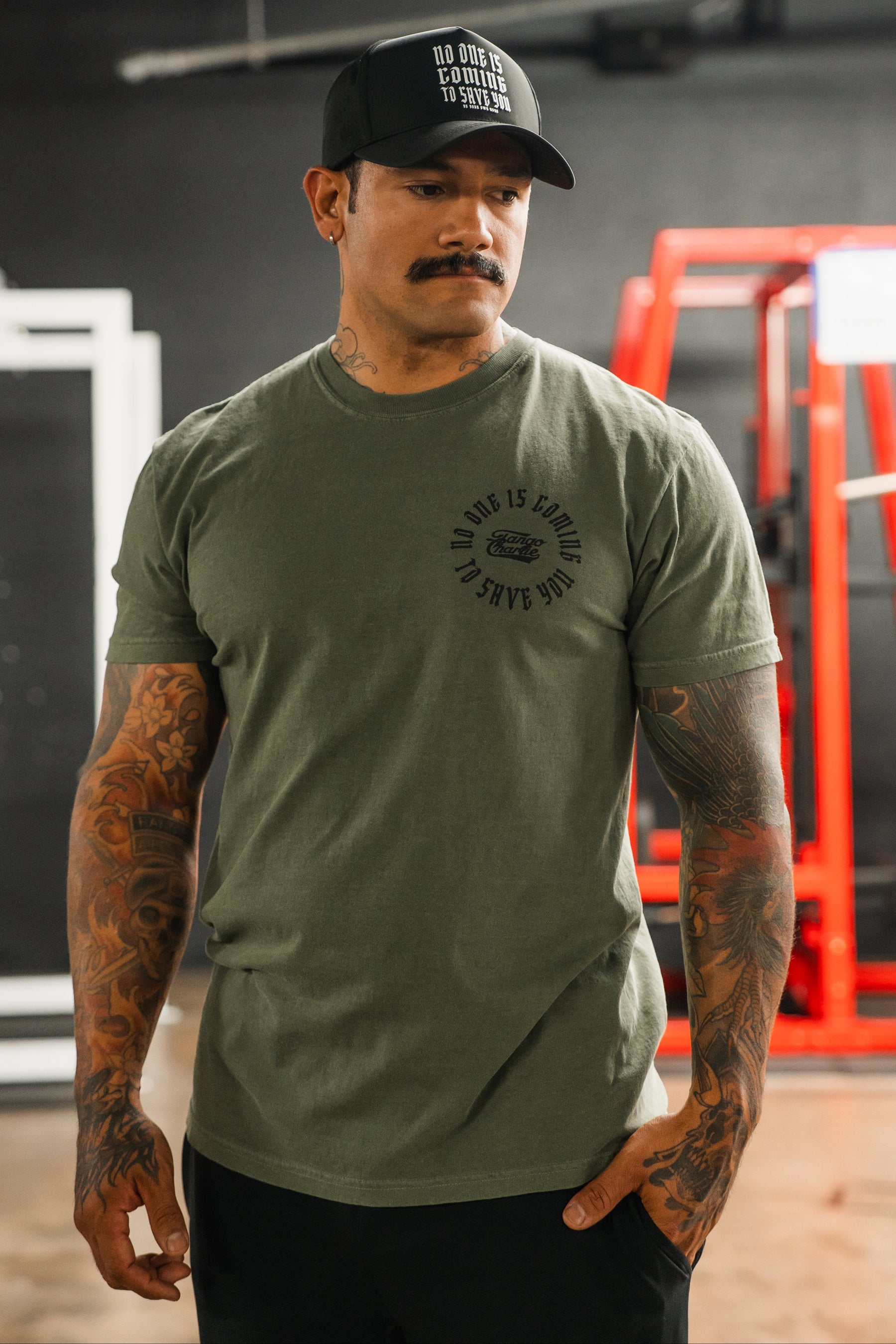 A tattooed man stands indoors wearing the olive green "No One Is Coming To Save You Hemp - Premium Tee" by Tango Charlie Apparel and a matching black cap. Red gym equipment is visible in the background.