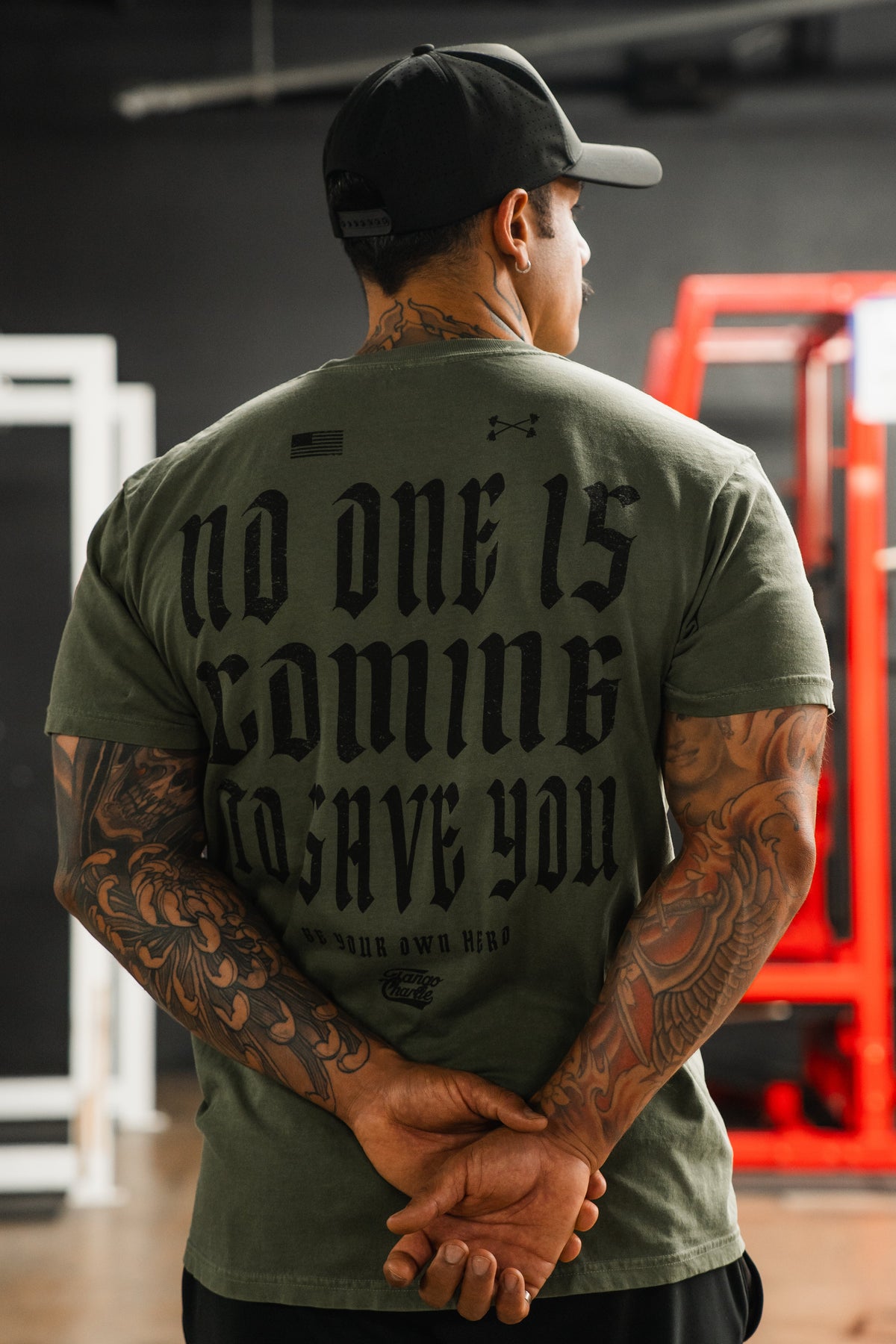 A tattooed man stands with hands behind his back wearing the No One Is Coming To Save You Hemp - Premium Tee by Tango Charlie Apparel. The green shirt features bold black text and gym equipment with a red frame can be seen in the background.
