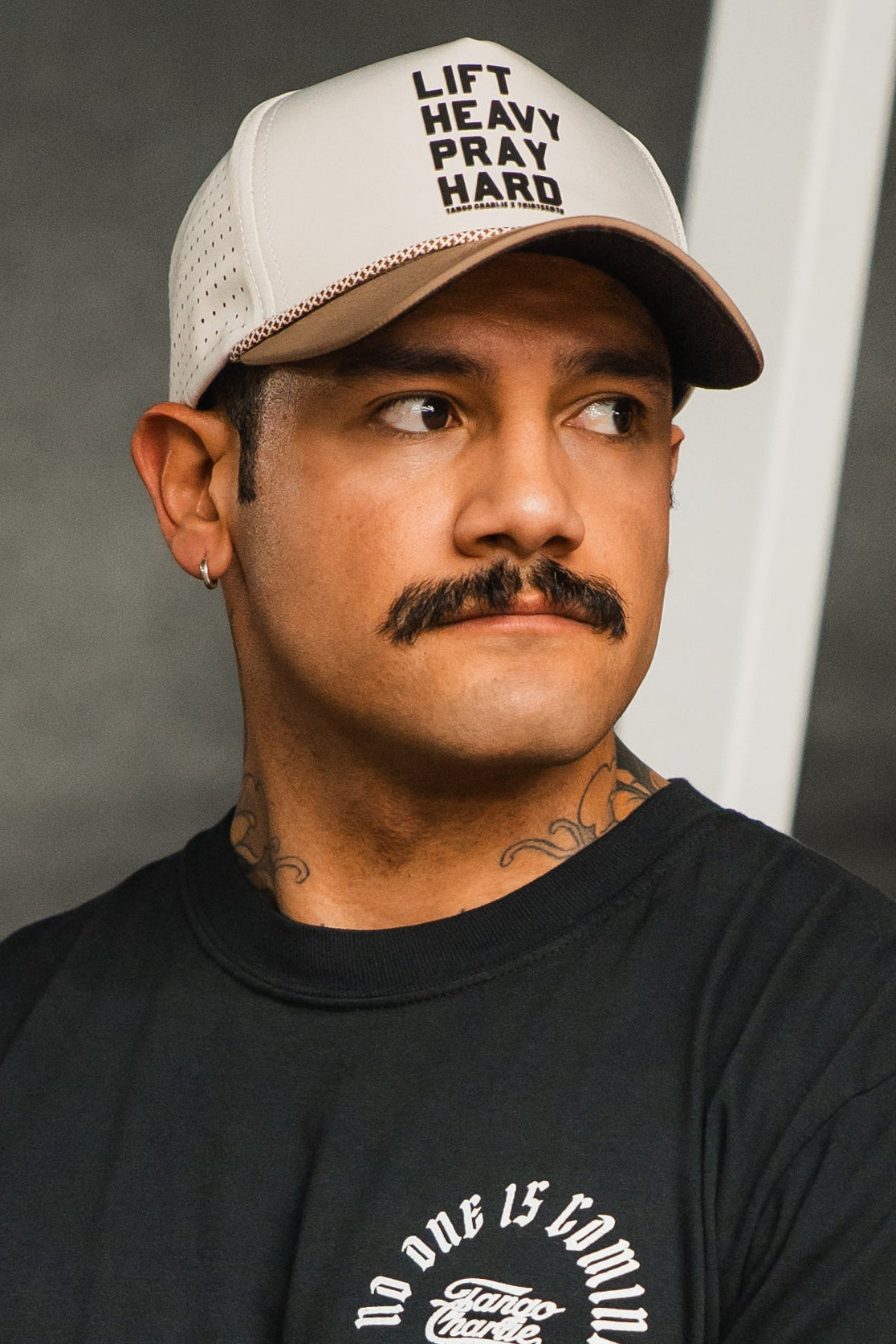 A man with a mustache, an earring, and a black t-shirt looks to the side while wearing the beige Lift Heavy. Pray Hard. Hydro - Snapback Hat by Tango Charlie Apparel against a blurred background.