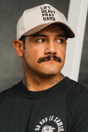 A man with a mustache, an earring, and a black t-shirt looks to the side while wearing the beige Lift Heavy. Pray Hard. Hydro - Snapback Hat by Tango Charlie Apparel against a blurred background.