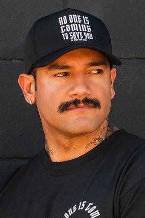 A man with a mustache and ear piercing wears the No One Is Coming To Save You Hydro Snapback Hat by Tango Charlie Apparel, featuring bold white gothic text, along with a black shirt. He stands against a dark wall, looking to his left.