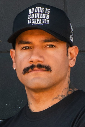 A man with a mustache and neck tattoos wears the Tango Charlie Apparel "No One Is Coming To Save You Hydro" snapback hat and a black shirt, standing before a dark background.