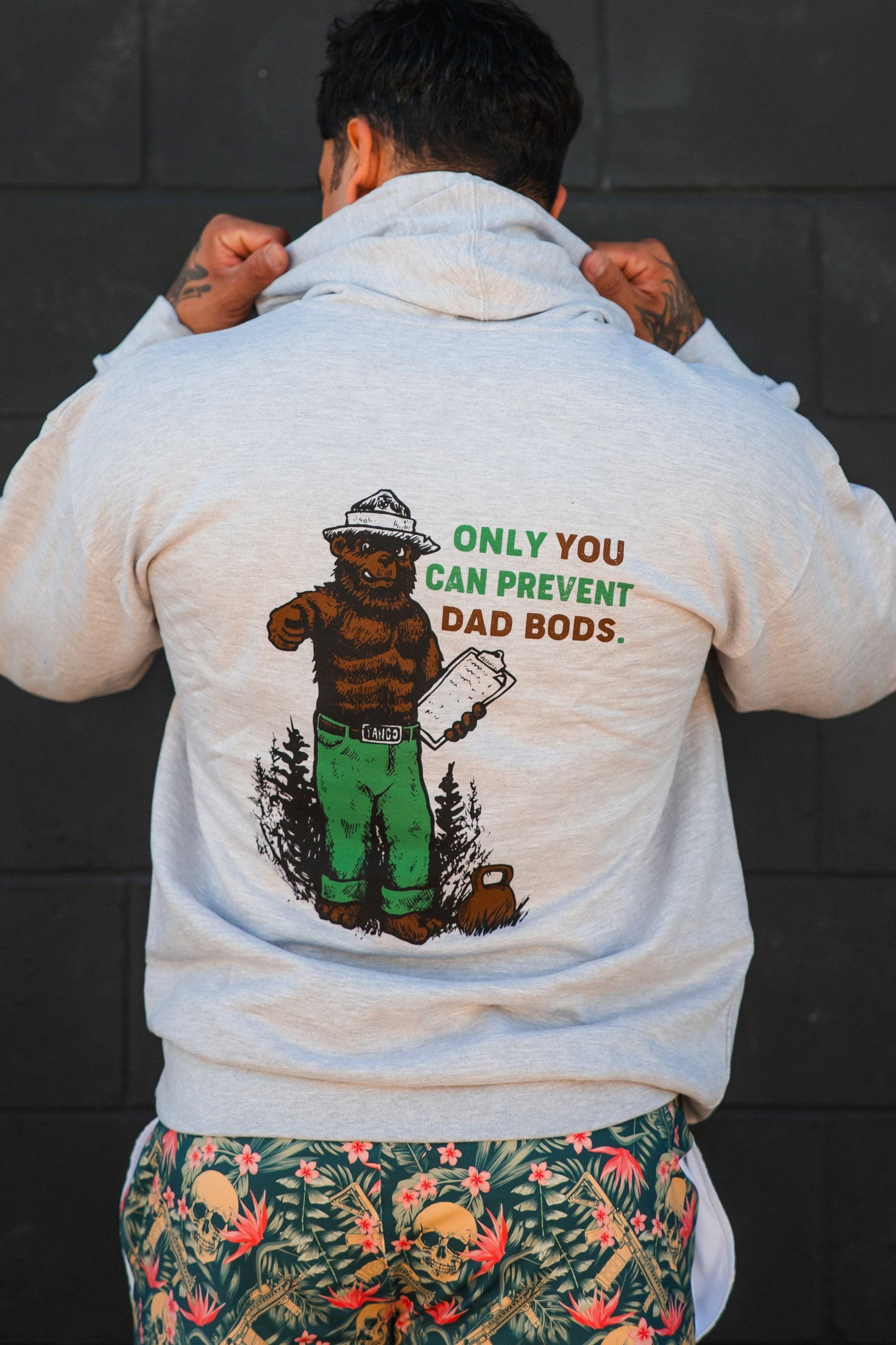 A person faces away from the camera, lifting the hood of Tango Charlie Apparel’s Only You Can Prevent Dad Bod Hoodie—gray with a bear illustration and humorous text—and wears colorful skull-patterned shorts.
