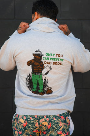 A person faces away from the camera, lifting the hood of Tango Charlie Apparel’s Only You Can Prevent Dad Bod Hoodie—gray with a bear illustration and humorous text—and wears colorful skull-patterned shorts.