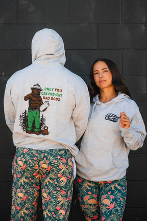 Two people in patterned leggings and gray Tango Charlie Apparel "Only You Can Prevent Dad Bod" hoodies stand by a dark wall. One shows the bear graphic with gym humor on the back; the other displays the front logo.