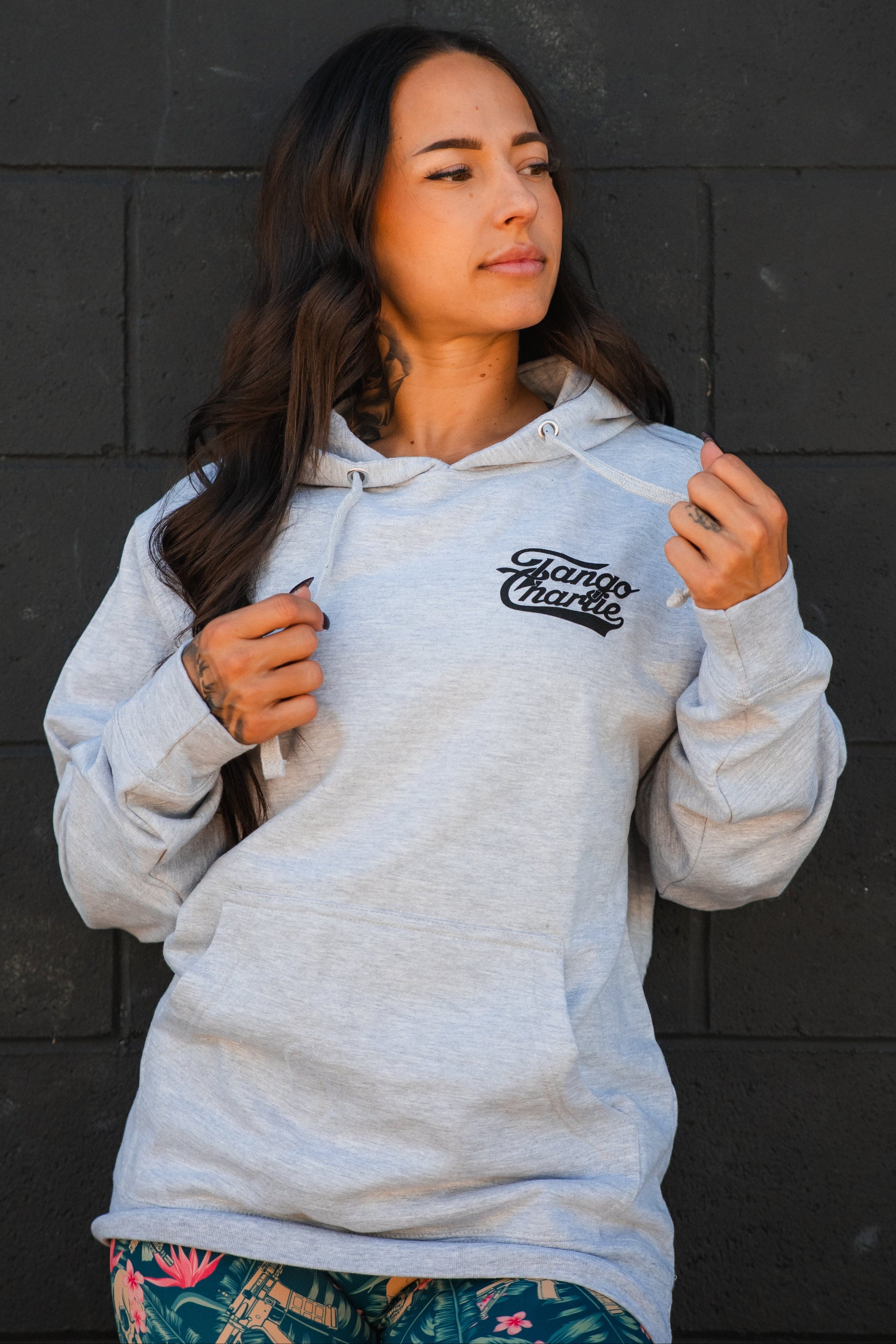 A woman with long dark hair models the Only You Can Prevent Dad Bod Hoodie by Tango Charlie Apparel, standing before a dark brick wall and holding the hoodie strings while looking to the side.