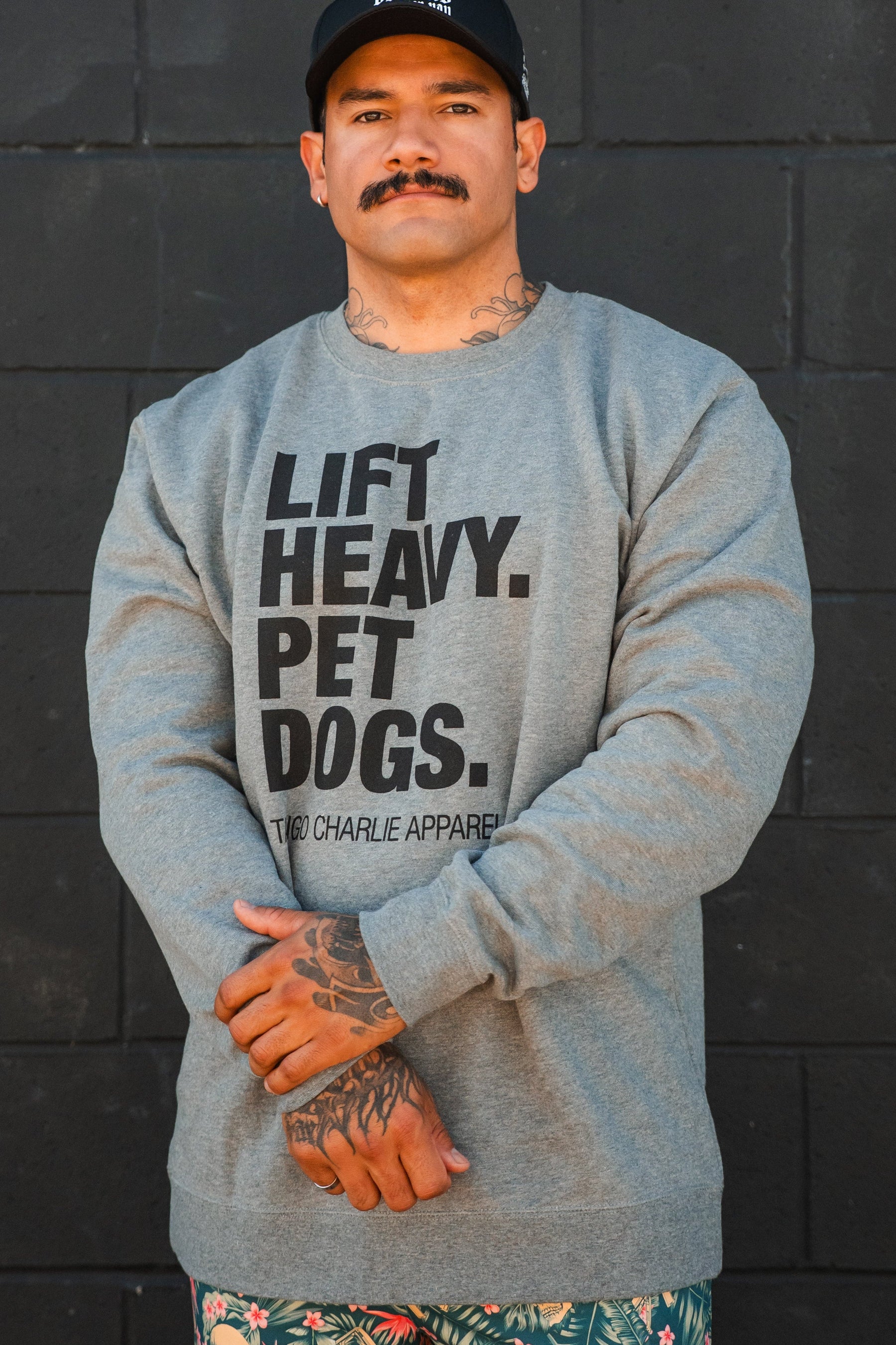 A man wearing the Lift Heavy, Pet Dogs Crewneck by Tango Charlie Apparel stands against a black brick wall, arms crossed. He sports a mustache, tattoos, a black cap, and patterned shorts, showing his love for dogs.