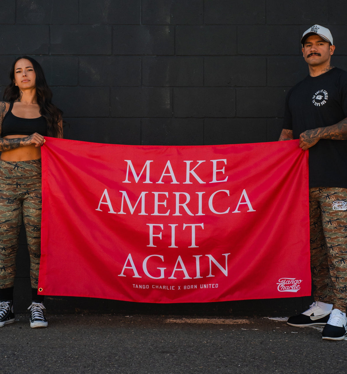 Two people in camo pants and black tops hold the Tango Charlie Apparel "Make America Fit Again" red polyester flag in front of a dark wall. The banner features bold white lettering.