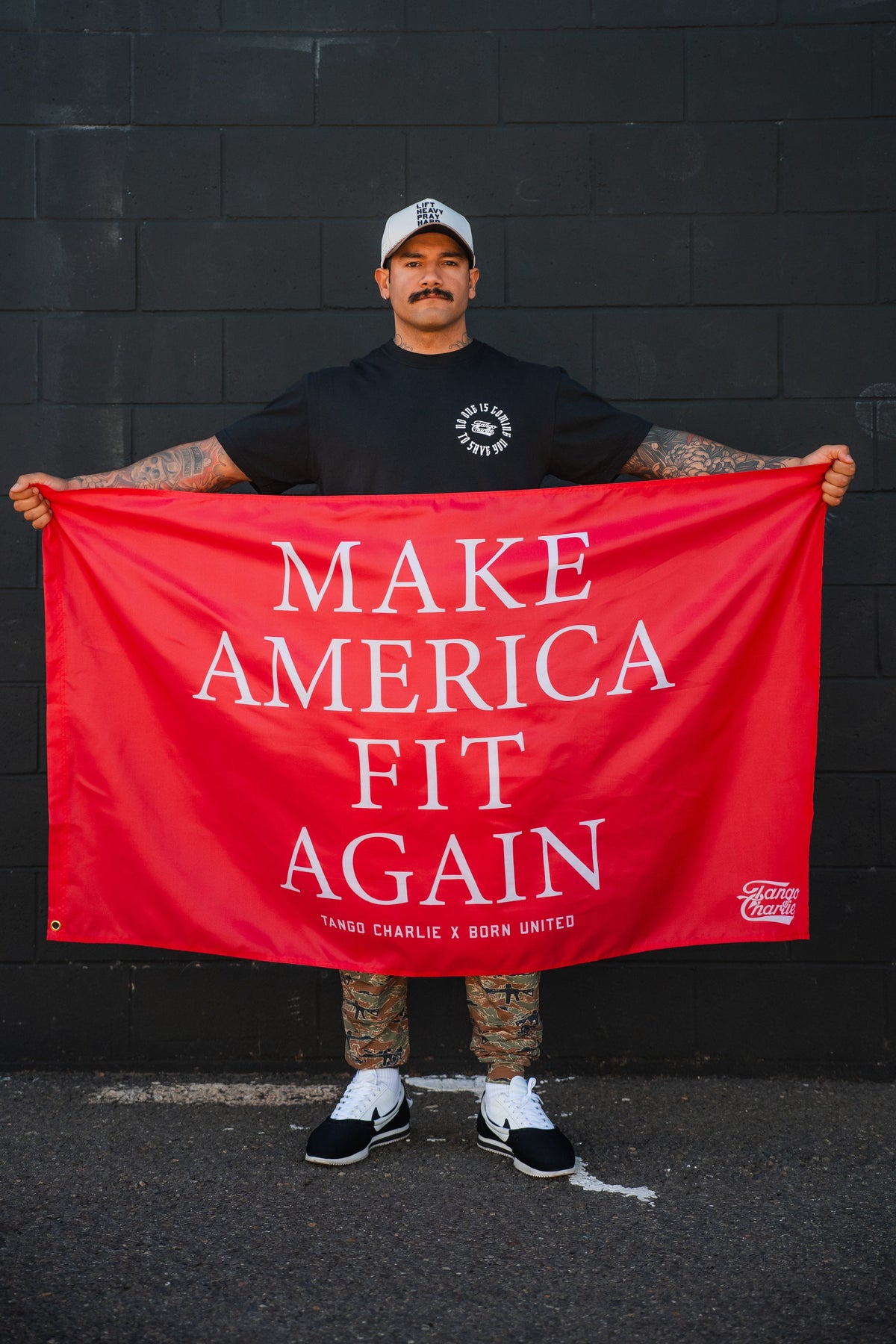 A tattooed man in a black t-shirt, white cap, and sneakers holds the Tango Charlie Apparel "Make America Fit Again" Flag against a black wall.