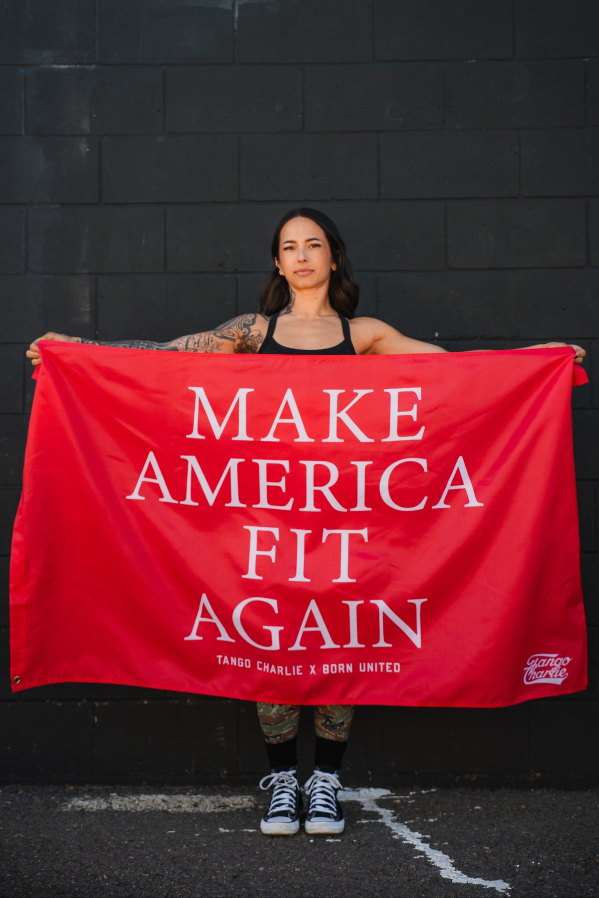 A tattooed woman stands before a black brick wall, holding the bold polyester "Make America Fit Again - Flag" by Tango Charlie Apparel.