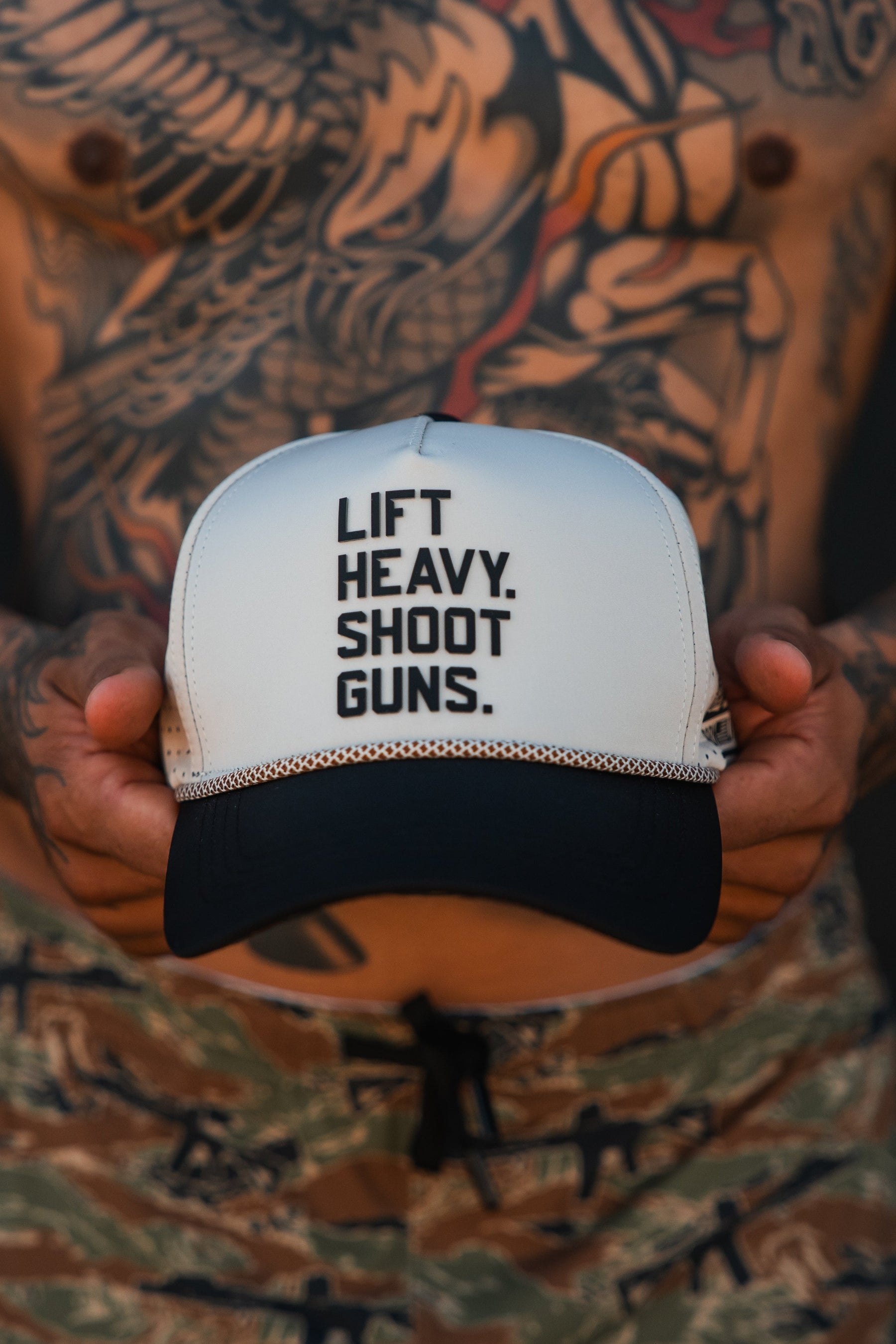 A shirtless person with arm and chest tattoos holds a Tango Charlie Apparel LHSG Hydro Snapback Hat that says LIFT HEAVY. SHOOT GUNS., while wearing camouflage pants.