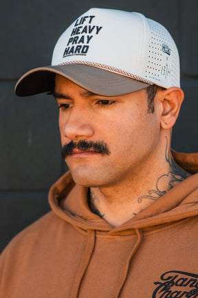 A man with a mustache, neck tattoos, and a tan hoodie looks down thoughtfully. He wears the Tango Charlie Apparel Lift Heavy. Pray Hard. Hydro Snapback Hat against a dark background.