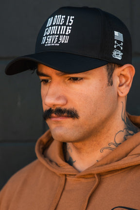 A man with a mustache and neck tattoos wears the Tango Charlie Apparel "No One Is Coming To Save You Hydro" snapback hat. The black, breathable cap features the slogan on the front. He pairs it with a brown hoodie against a dark background.
