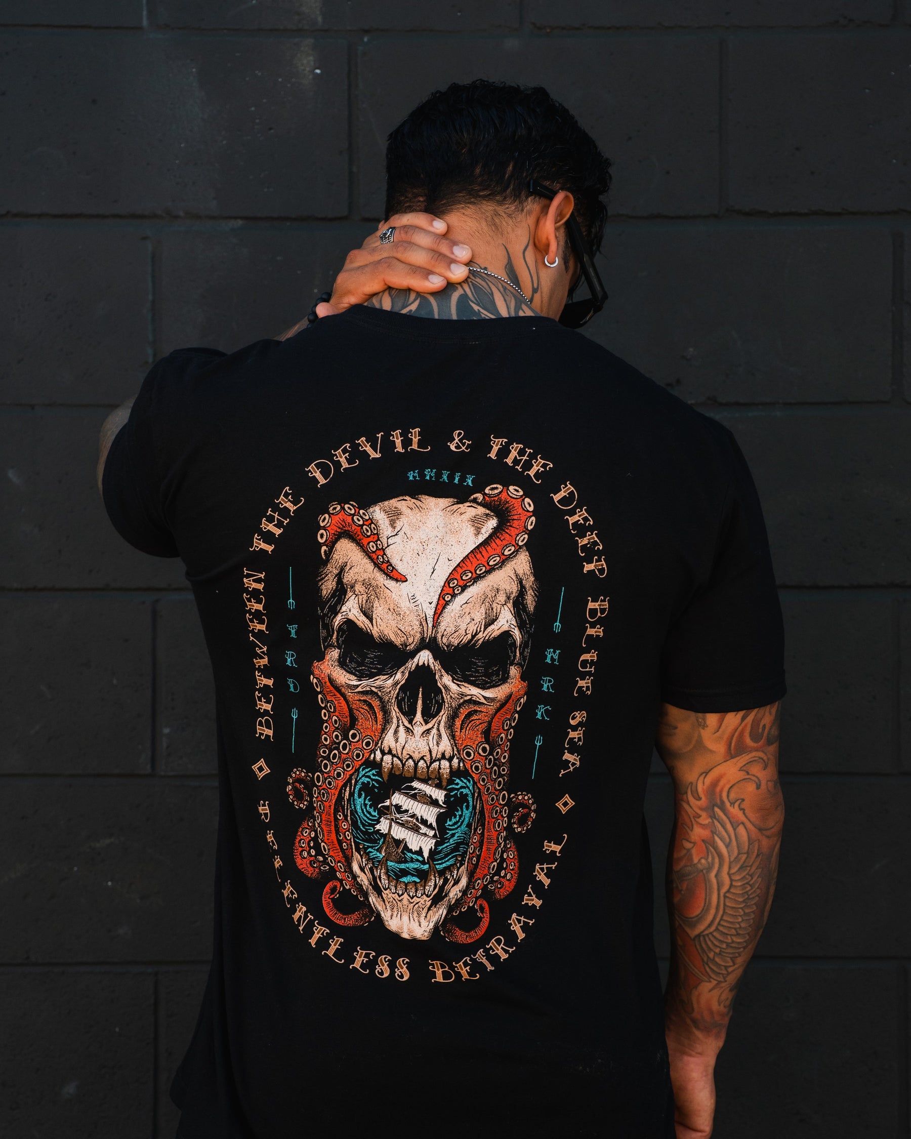 A tattooed person faces away, wearing the "Between the Devil & The Deep Blue Sea T-Shirt" by Relentless Betrayal, featuring a bold skull and snake design with the text: “Between the Devil & The Deep Blue Sea Relentless Betrayal.”.