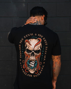A tattooed person faces away, wearing the "Between the Devil & The Deep Blue Sea T-Shirt" by Relentless Betrayal, featuring a bold skull and snake design with the text: “Between the Devil & The Deep Blue Sea Relentless Betrayal.”.