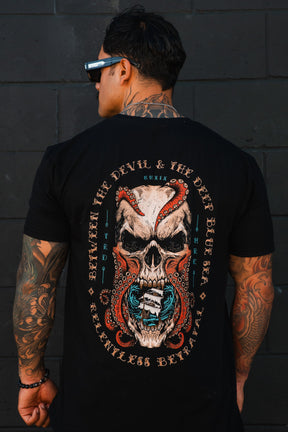 A tattooed man in sunglasses turns away, showcasing the Relentless Betrayal "Between the Devil & The Deep Blue Sea" T-Shirt with a bold skull, octopus, and jaws graphic surrounded by the statement text.
