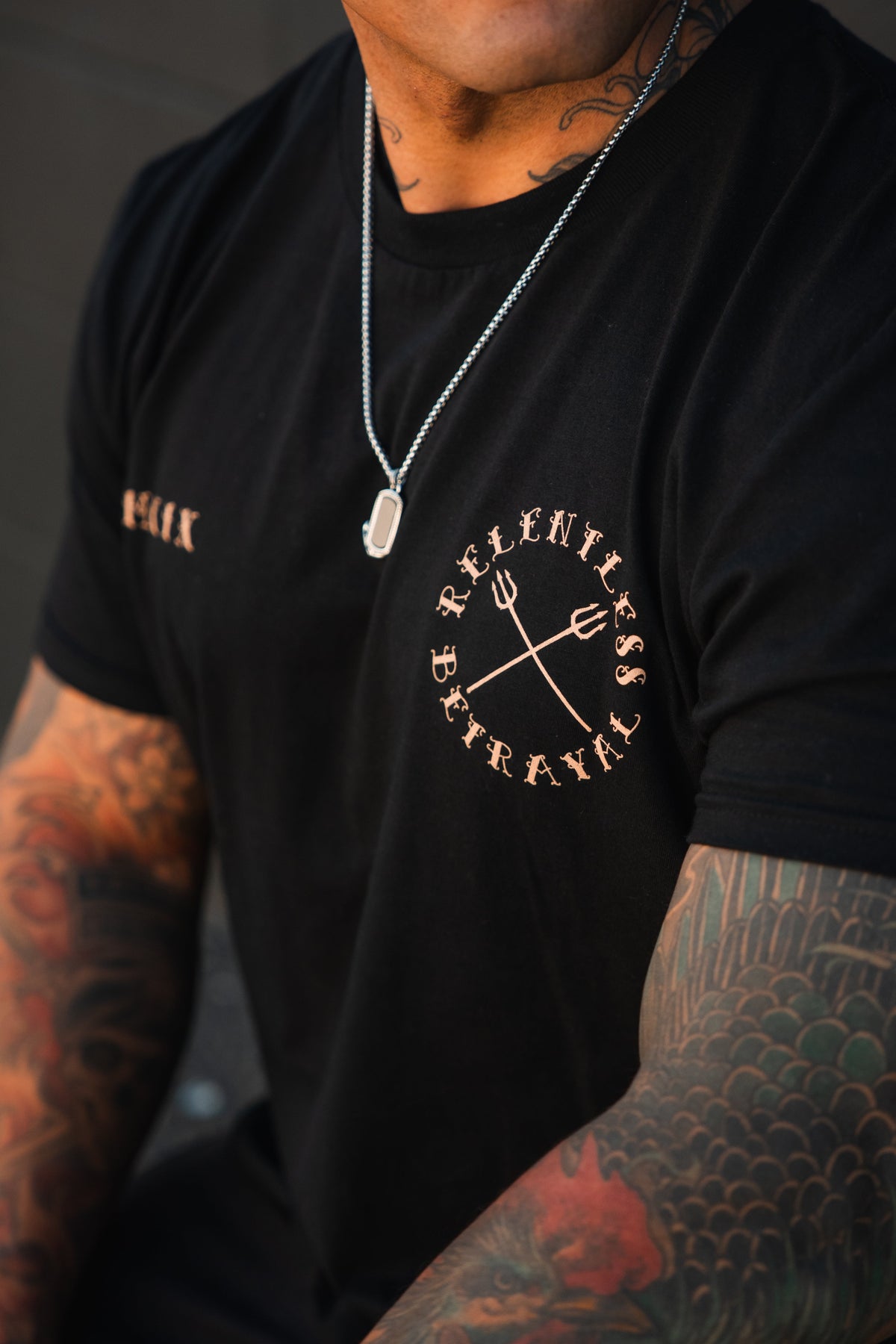 A tattooed individual wears the "Between the Devil & The Deep Blue Sea" unisex t-shirt by Relentless Betrayal, showcasing its circular graphic with crossed pitchforks, paired with a silver chain necklace and rectangular pendant.