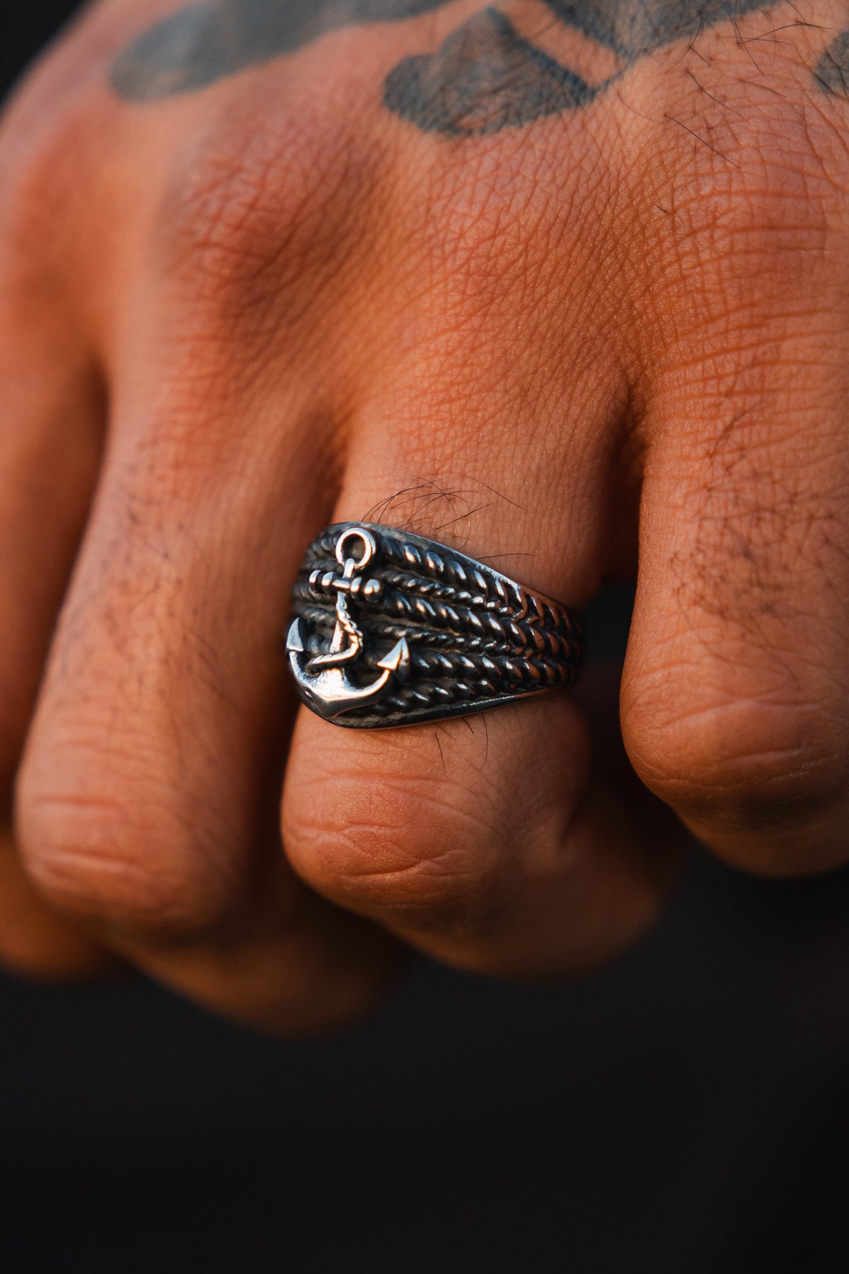 A close-up of a hand wearing The Gravewater Ring by Relentless Betrayal—this handcrafted, silver-toned stainless steel ring features anchor and rope details, resting above textured skin with a visible knuckle tattoo.