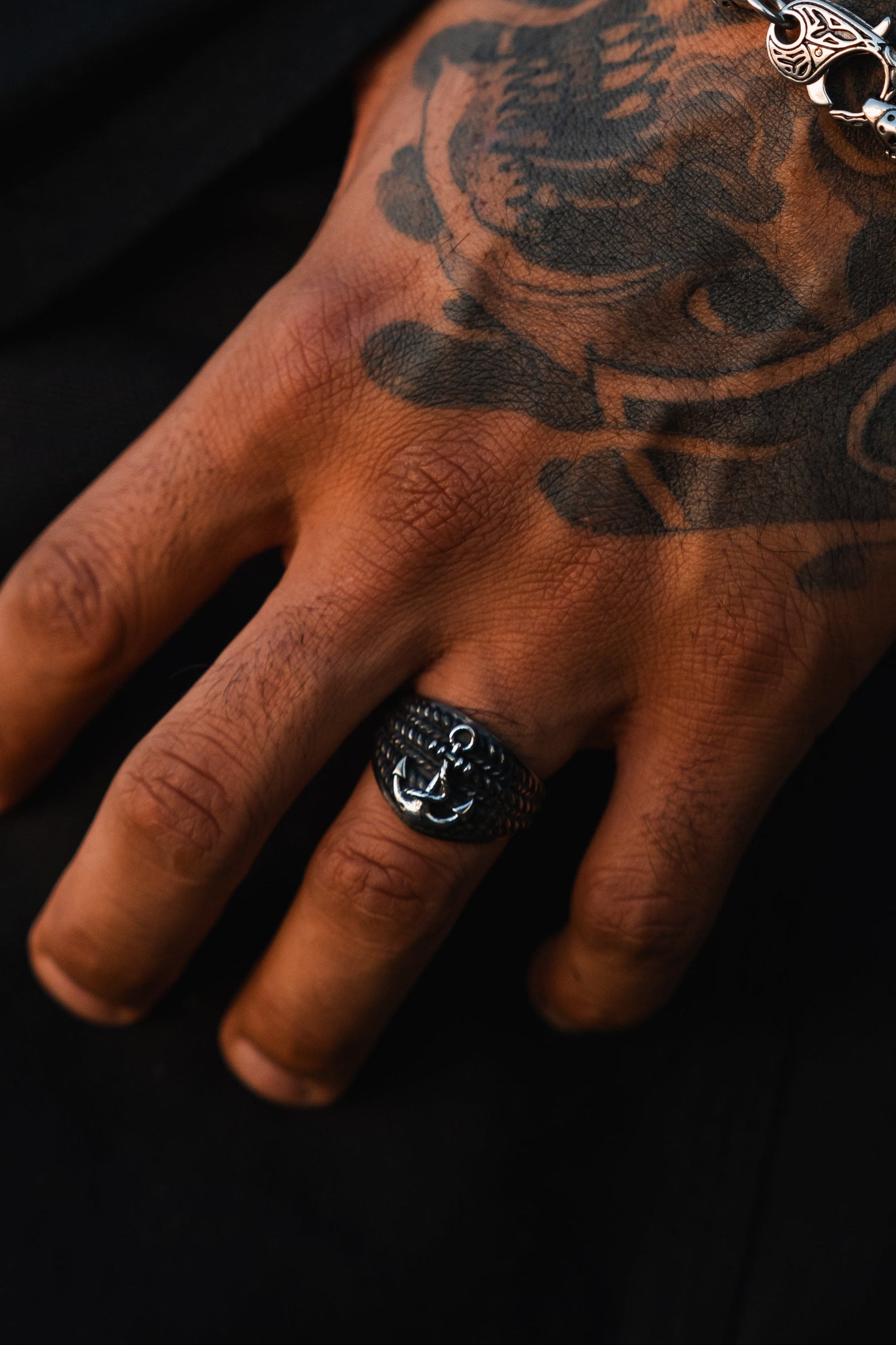 A close-up of a tattooed hand wears The Gravewater Ring by Relentless Betrayal—a handcrafted stainless steel ring featuring a silver anchor—on the middle finger, paired with a decorative chain bracelet against a dark background.