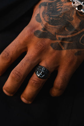 A close-up of a tattooed hand wears The Gravewater Ring by Relentless Betrayal—a handcrafted stainless steel ring featuring a silver anchor—on the middle finger, paired with a decorative chain bracelet against a dark background.