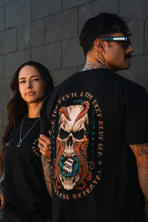A man and woman in black stand by a dark brick wall. The man shows off the back of his Relentless Betrayal "Between the Devil & The Deep Blue Sea" T-shirt, featuring a bold skull and snake graphic. Both have tattoos.