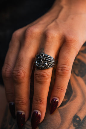 A hand with dark red, pointed nails wears Relentless Betrayal’s handcrafted Gravewater Ring—stainless steel with an anchor design—resting on tattooed skin, lighting accentuating the details of the ring and tattoo.