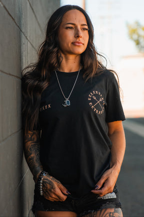 A woman with long brown hair, tattoos, and a serious expression stands outdoors in black shorts and a necklace, wearing the Relentless Betrayal "Between the Devil & The Deep Blue Sea" unisex t-shirt by a concrete wall.