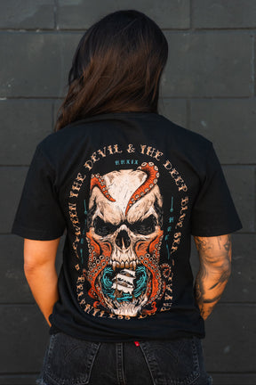 A person with long hair and a tattooed arm wears the Relentless Betrayal "Between the Devil & The Deep Blue Sea" unisex T-shirt, featuring a large skull with red tentacles and blue accents, standing by a dark gray brick wall.