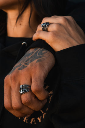 Close-up of tattooed hands wearing silver rings, including Relentless Betrayal's handcrafted Gravewater Ring and a detailed anchor ring. The person is dressed in a black hoodie.