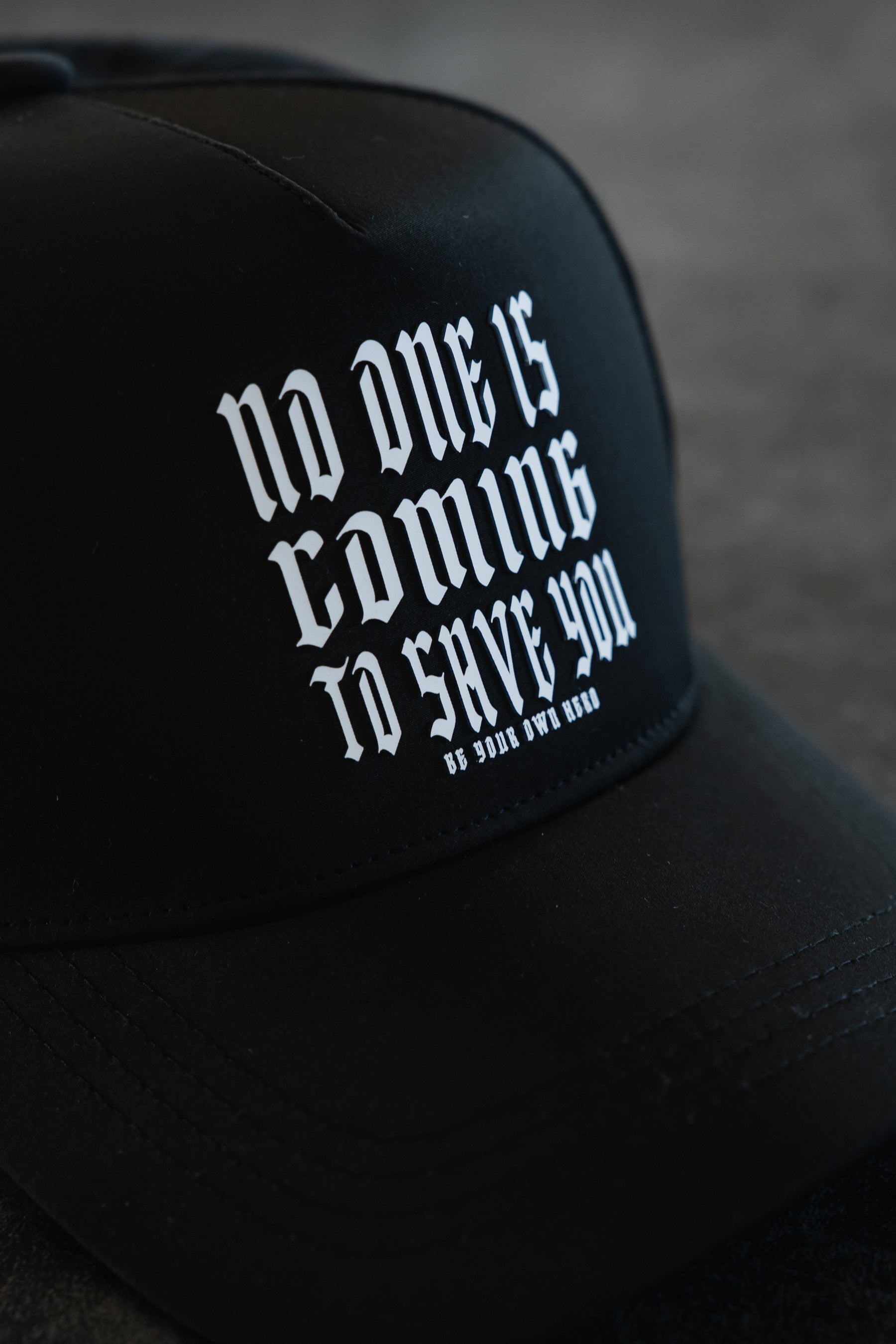 A close-up of Tango Charlie Apparel’s No One Is Coming To Save You Hydro Snapback Hat, featuring black hydro-resistant fabric and white gothic-style text: “No one is coming to save you. Be your own hero.”.