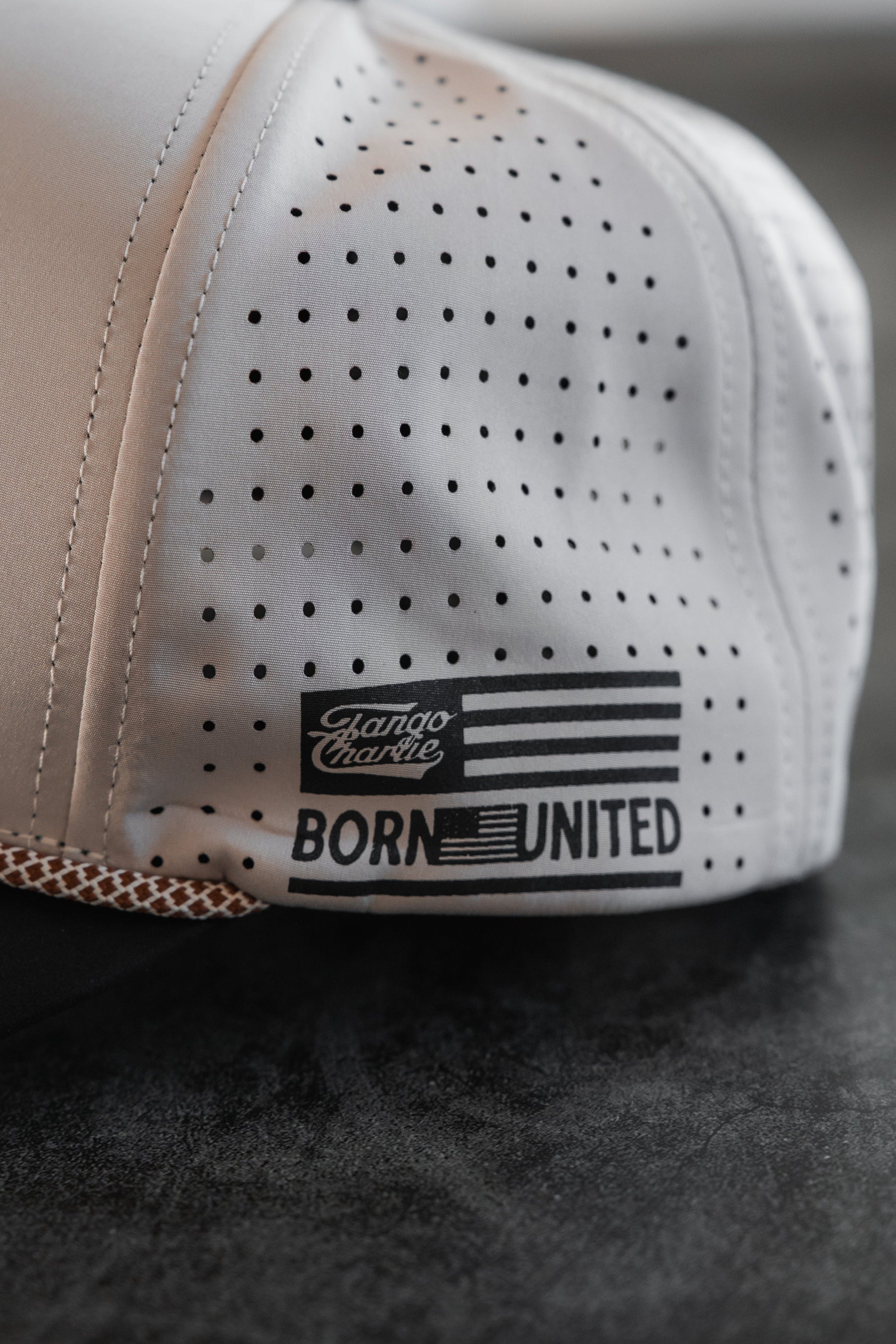 A close-up of the LHSG Hydro - Snapback Hat by Tango Charlie Apparel, featuring black dotted details and a BORN UNITED patch with an American flag and Canoe Charlie logo, from their collaboration, resting on a dark surface.