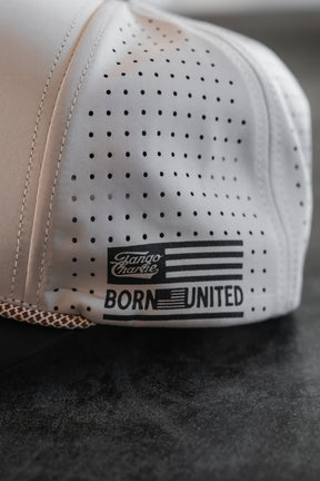 A close-up of the LHSG Hydro - Snapback Hat by Tango Charlie Apparel, featuring black dotted details and a BORN UNITED patch with an American flag and Canoe Charlie logo, from their collaboration, resting on a dark surface.