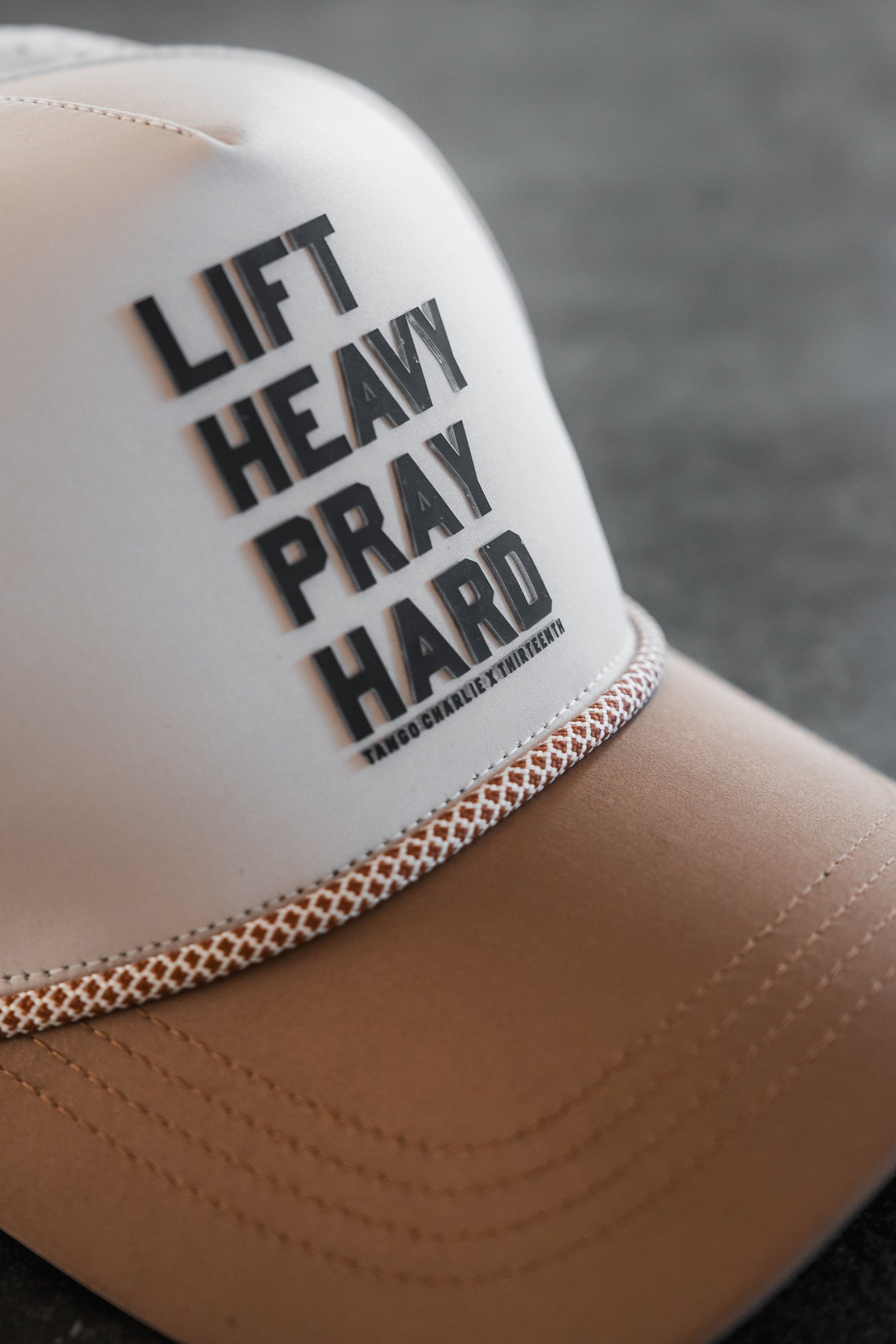 The Lift Heavy. Pray Hard. Hydro - Snapback Hat by Tango Charlie Apparel features bold black text on a beige and tan cap with decorative stitching above the brim, shown resting on a dark, textured surface.