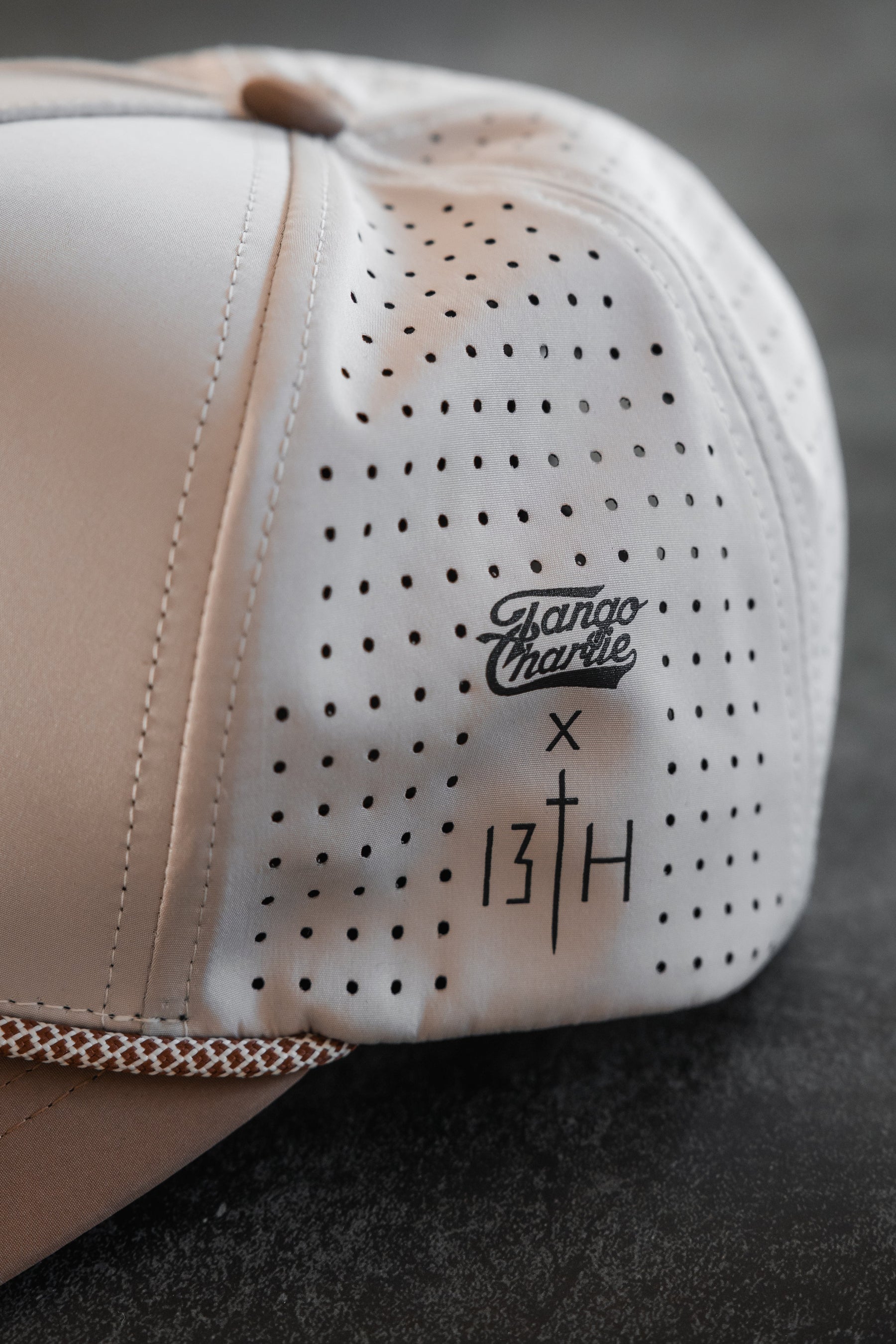 Close-up of the Lift Heavy. Pray Hard. Hydro Snapback Hat by Tango Charlie Apparel, in light beige with water resistance, small perforated holes, and black printed logos on a dark textured surface.