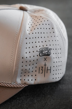 Close-up of the Lift Heavy. Pray Hard. Hydro Snapback Hat by Tango Charlie Apparel, in light beige with water resistance, small perforated holes, and black printed logos on a dark textured surface.