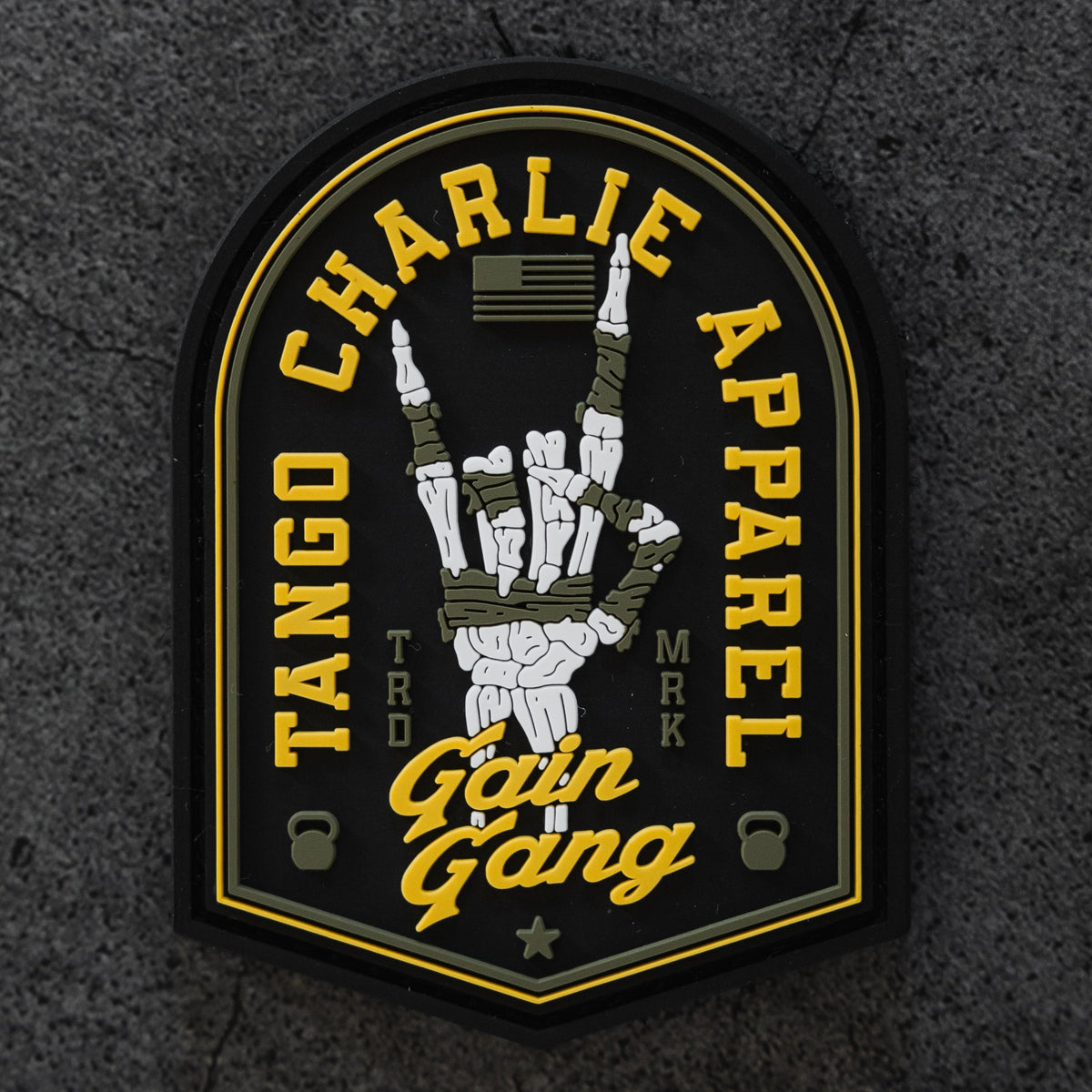 A 3-inch Velcro Gain Gang - Patch by Tango Charlie Apparel features a skeleton hand rock sign, yellow brand text, Gain Gang in script, small kettlebells, and an American flag on a gray textured background.