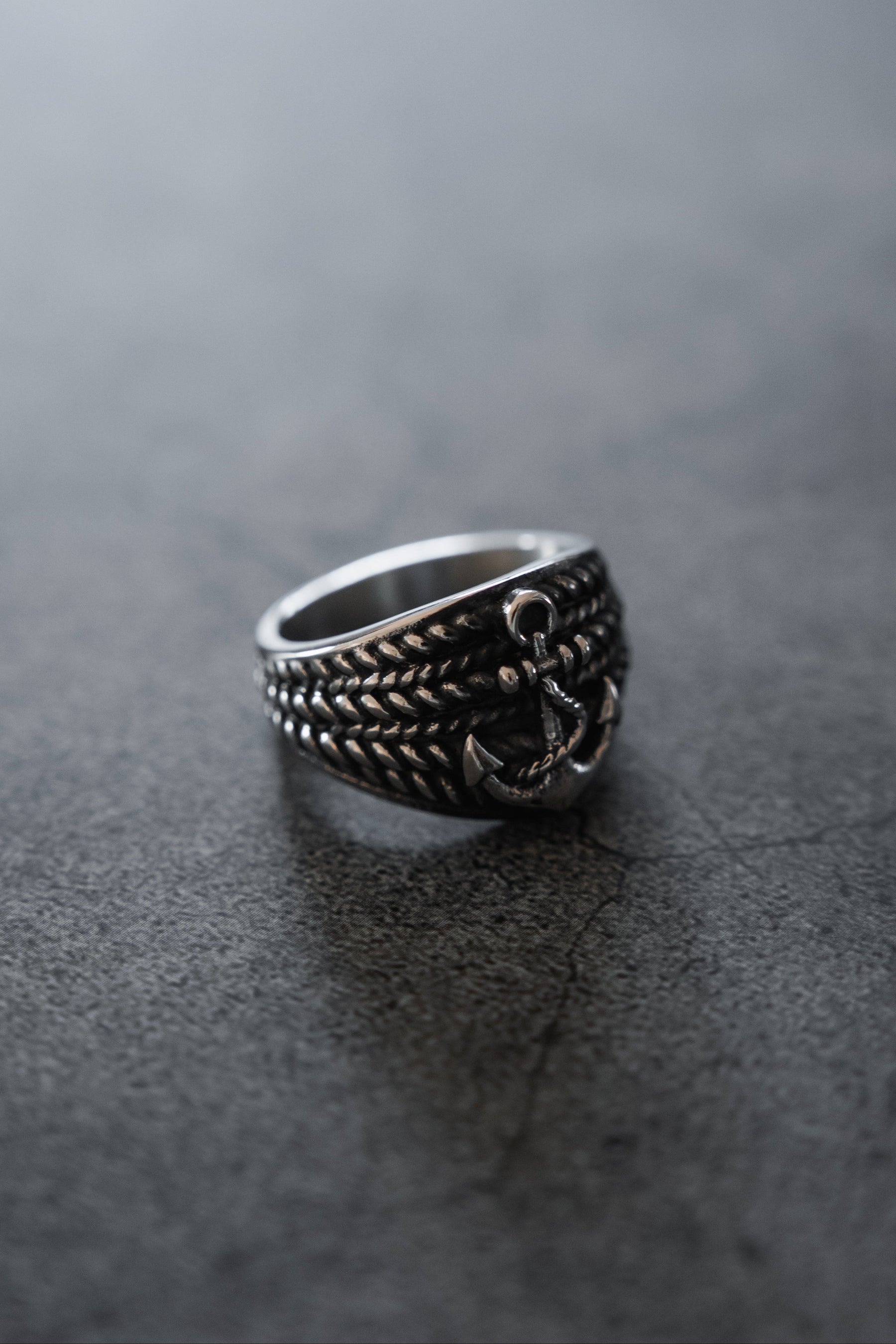 The Gravewater Ring by Relentless Betrayal features a handcrafted textured band and anchor design in silver stainless steel, displayed on a smooth dark gray surface.