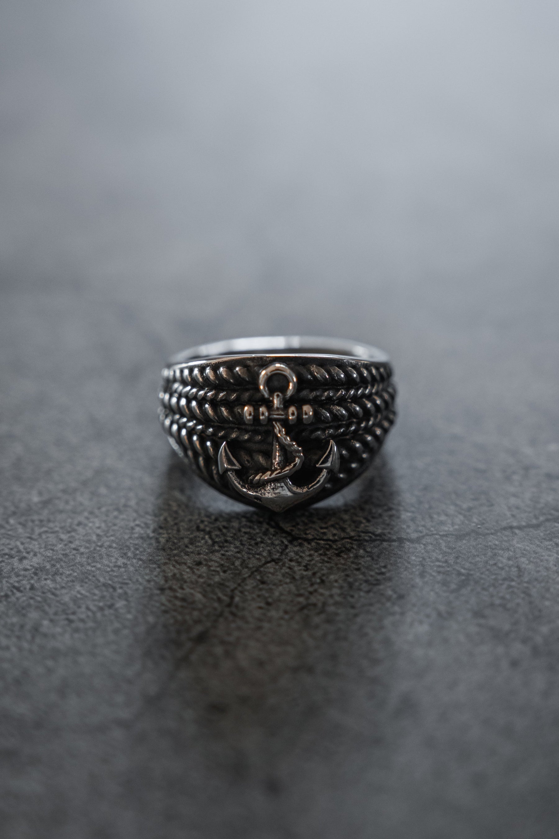The Gravewater Ring by Relentless Betrayal, featuring a handcrafted raised anchor and rope-like detailing, rests on a smooth dark gray surface.