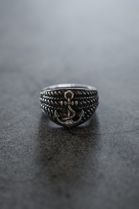 The Gravewater Ring by Relentless Betrayal, featuring a handcrafted raised anchor and rope-like detailing, rests on a smooth dark gray surface.