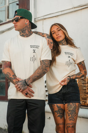 Two tattooed individuals stand side by side, proudly wearing matching Tattooed Child of God Premium Ivory T-Shirts from Relentless Betrayal’s Christian apparel collection. Each shirt showcases a striking “Tattooed Child of God” graphic, blending faith and style effortlessly as they pose in front of a building with white walls and windows.